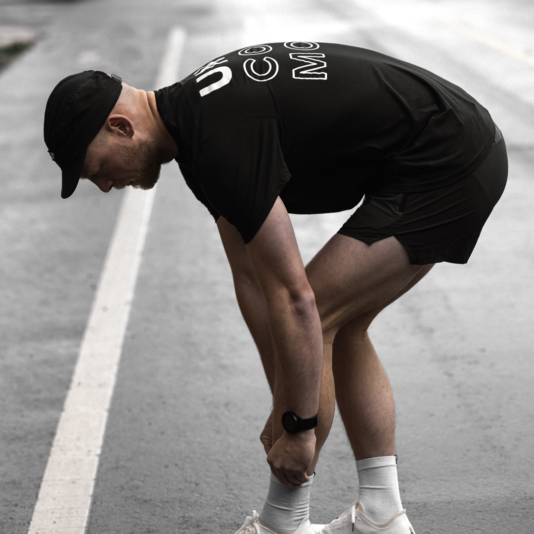 A man bending over and tying up the laces on his white running shoes, wearing a black running cap, t-shirt which says "Uncommon" on the back and split shorts.