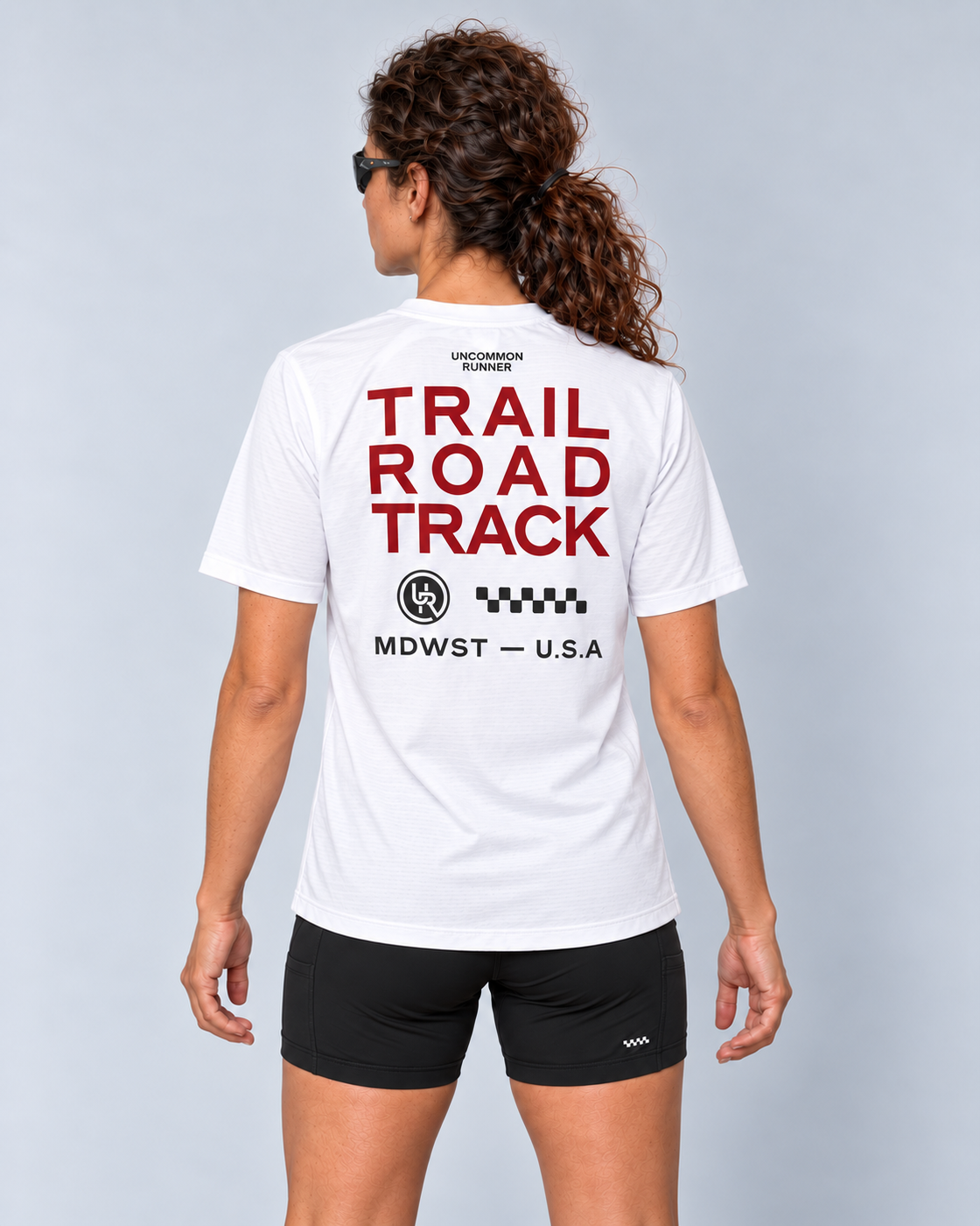 Back studio view of a female model wearing the white Three Ways short-sleeve TempoTech running tee with red Trail Road Track lettering and charcoal checker artwork and black Shift-5 shorts