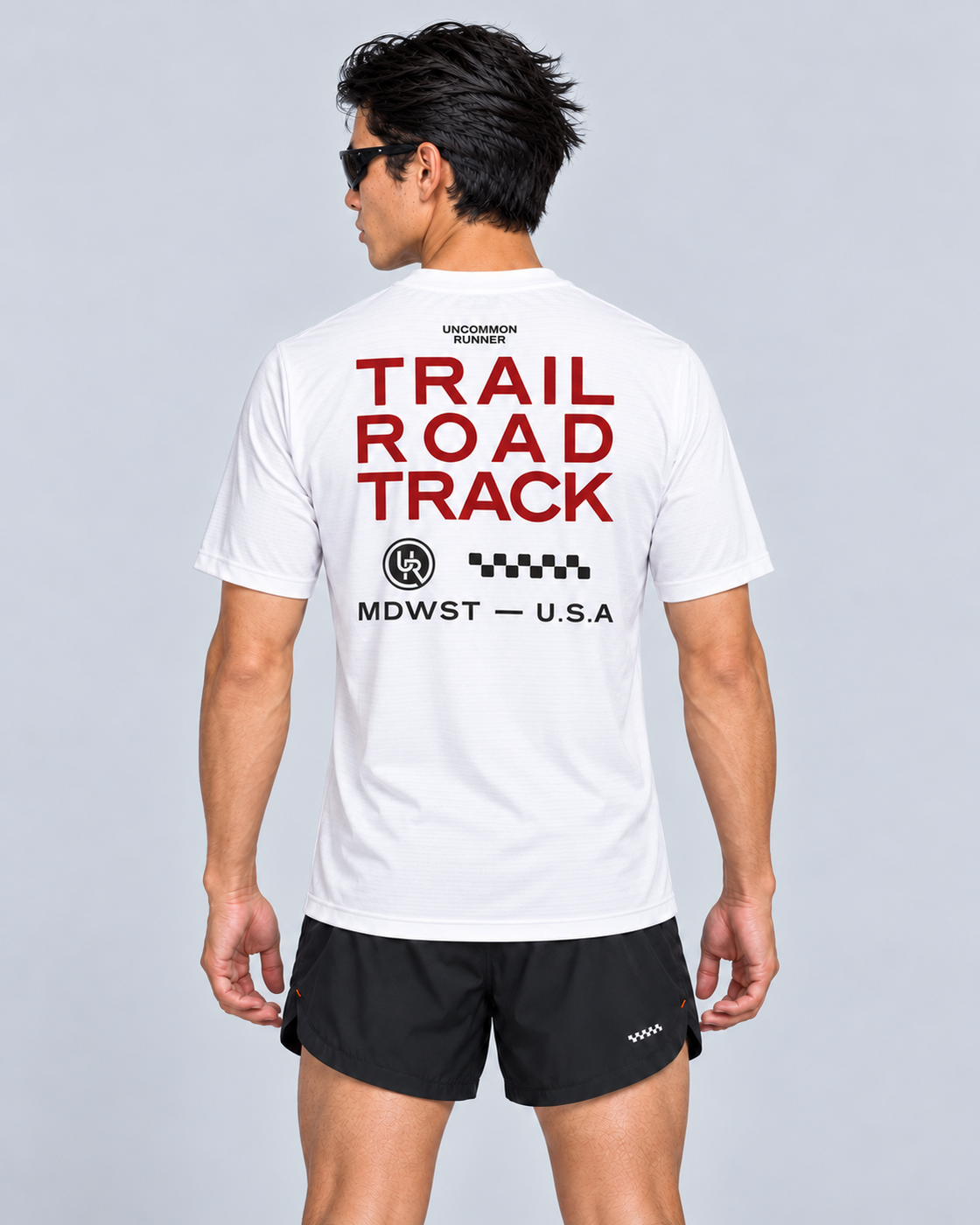 Back studio view of a male model wearing the white Three Ways short-sleeve TempoTech running tee with red Trail Road Track lettering and charcoal checker artwork and black Phanto-3 race shorts