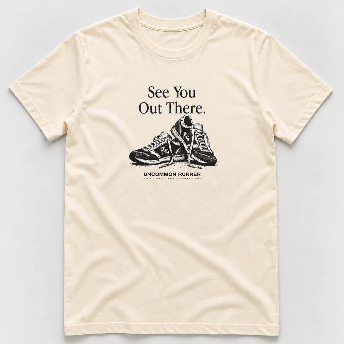 Front mockup of Uncommon Runner Kicks cotton tee in ecru, with See You Out There text and black-and-white shoe graphic.