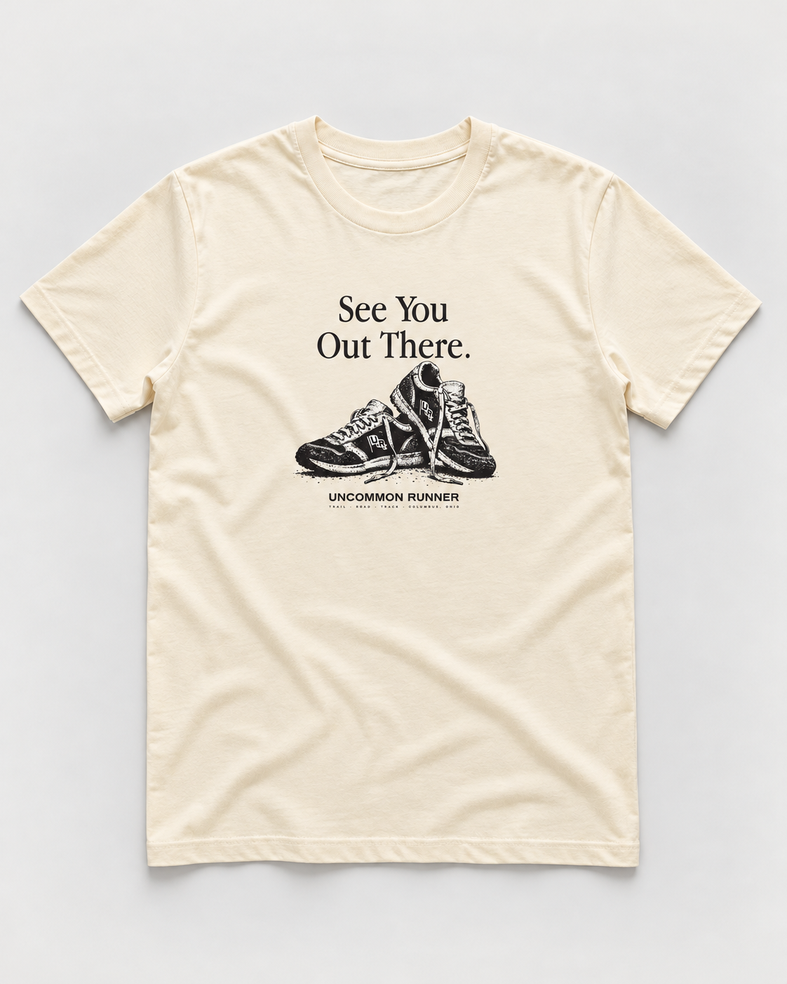 Front mockup of Uncommon Runner Kicks cotton tee in ecru, with See You Out There text and black-and-white shoe graphic.